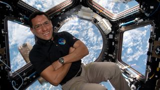 A man in khakis crosses his arms as he floats in front of a hexagonal window that looks down at Earth from orbit. 