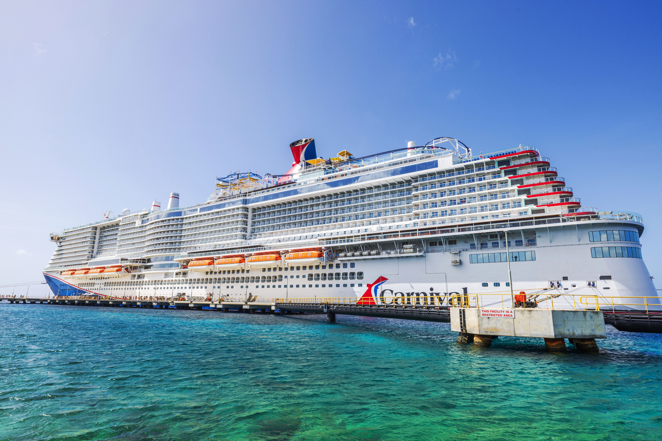 Carnival cruise ship docked in turquoise water Caribbean sea under clear blue sky with lifeboats along white hull near pier. 