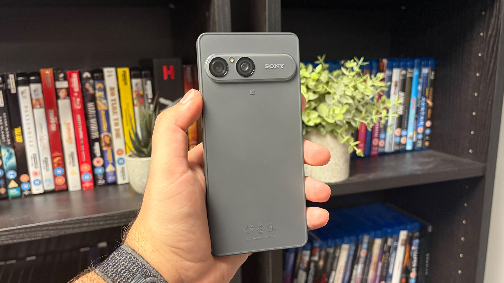 Sony Xperia 10 VIII smartphone held in hand in front of bookcase with DVDs, showing back of device