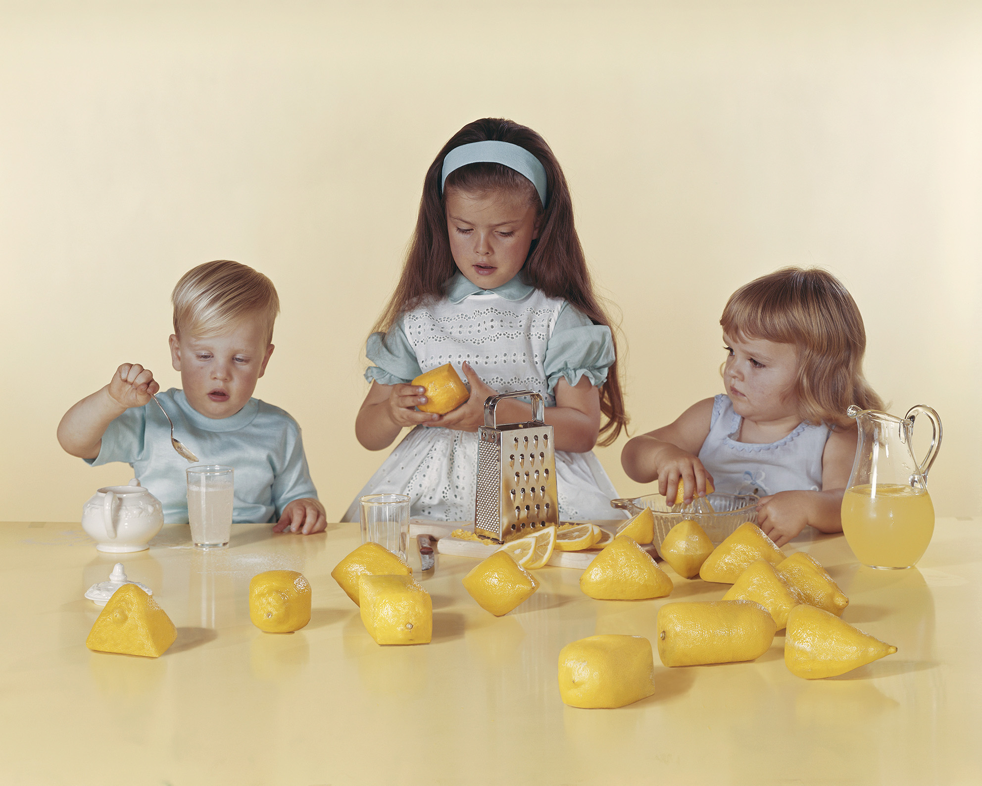 Colour photograph of three children sat at a table with lemons spread out, along with a grater and a jug