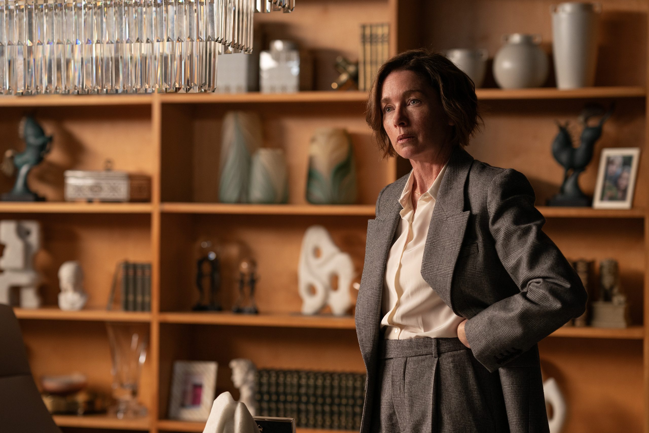 Julianne Nicholson, standing next to a chandelier in front of a wall of shelves and knick-knacks, in 'Paradise' season 1.