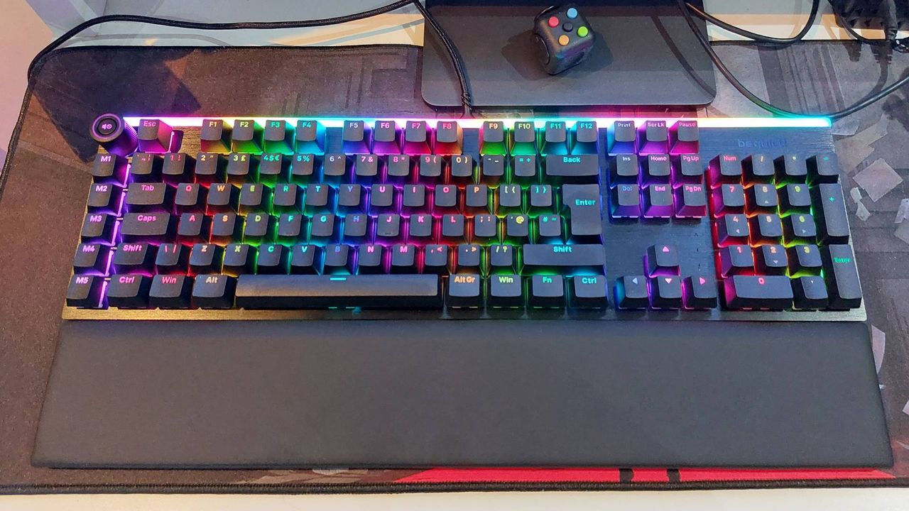 Be Quiet's Light Mount mechanical gaming keyboard sits on a red and black mouse mat. The magnetic wrist rest is attached and the RGB lights are on.