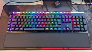 Be Quiet's Light Mount mechanical gaming keyboard sits on a red and black mouse mat. The magnetic wrist rest is attached and the RGB lights are on.