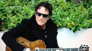 Neal Schon in sunglasses, sitting in front of a bush holding a guitar