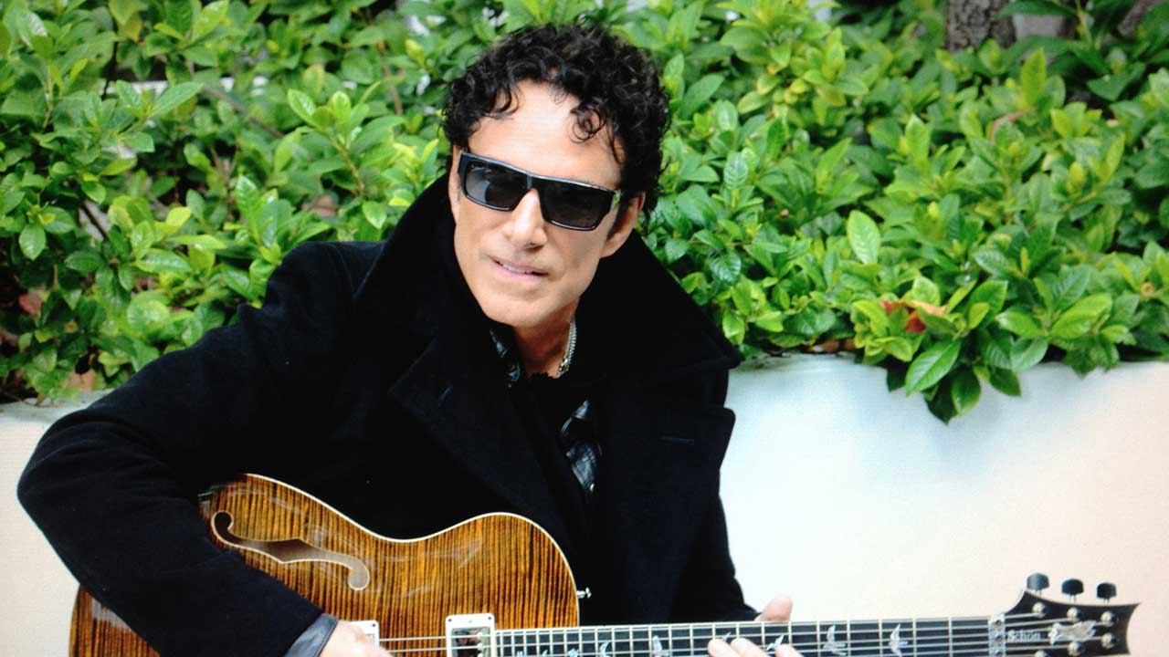 Neal Schon in sunglasses, sitting in front of a bush holding a guitar
