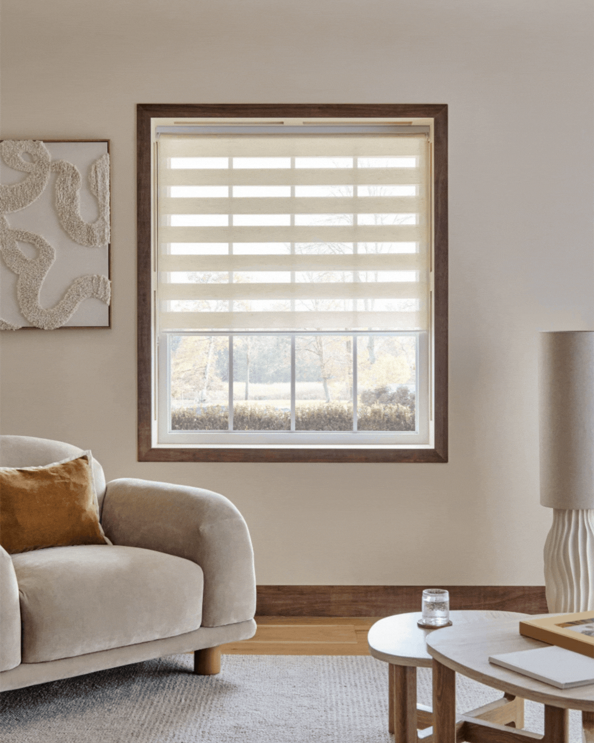 GIF of two rotating images that show cream day and night blinds on a window in a neutral living room. The walls are beige with wooden trim. There is a sand colored, velvet accent chair.