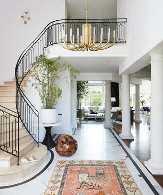 An entryway with curved black railing staircase, white columns, brass chandelier and open-plan access to the kitchen