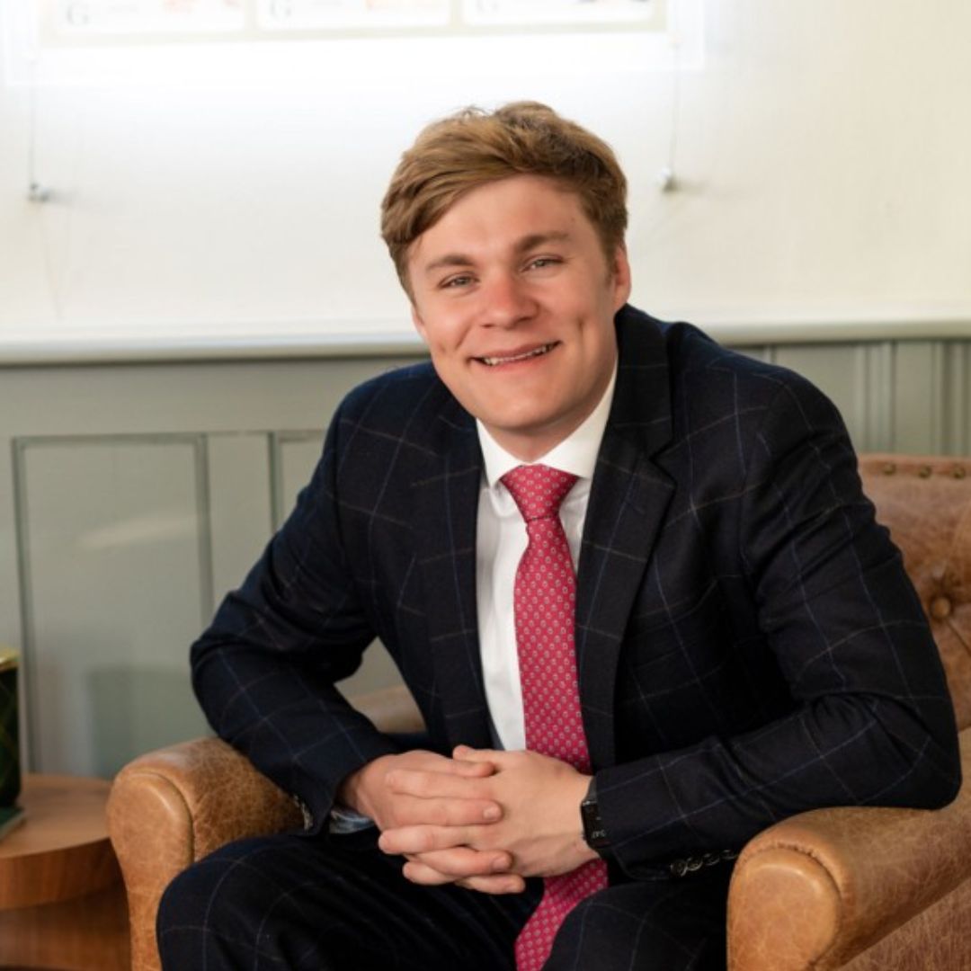 A smiling blonde man sitting in a leather armchair wearing a suit with a red tie