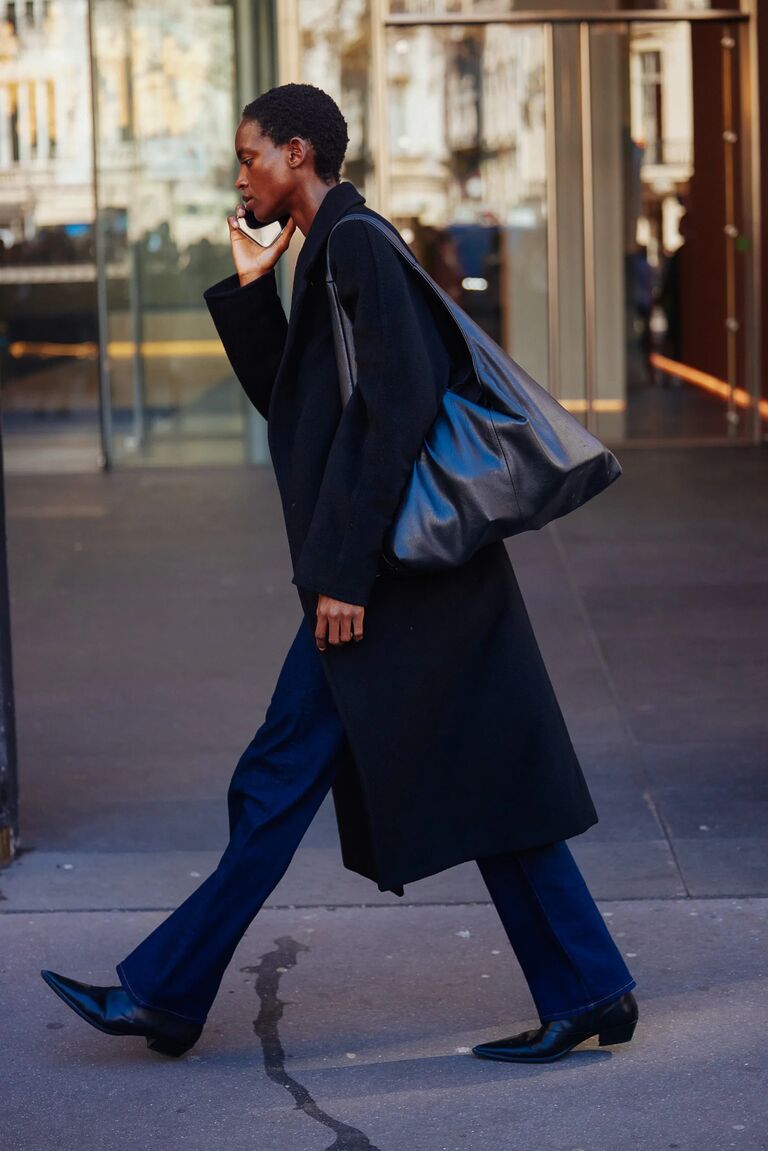 A women wears a black coat, blue bootcut jeans and black boots at Paris fashion week