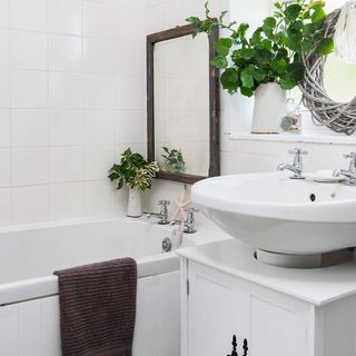 a bathroom with a white bath, white sink and unit and white tiles, with a mirror with a brown frame above the taps on the bath and two pot plants on the bath and by the window