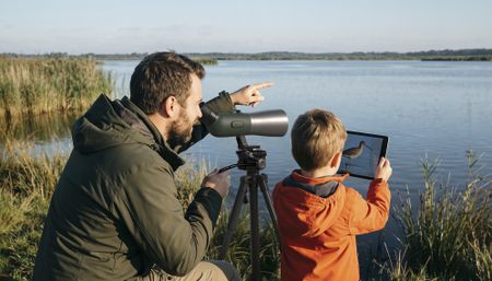 Man and child looking into an AI-powered spotting scope by the side of a lake, identifying a distant bird