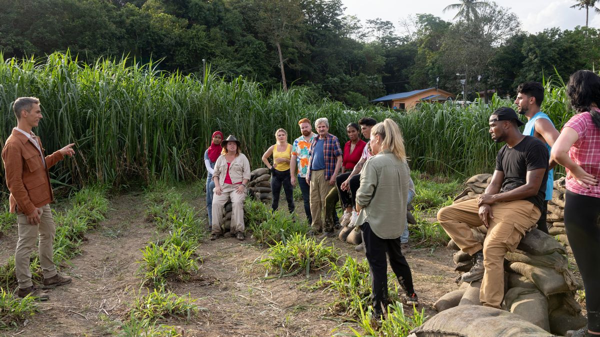 The Mole season 2 host Ari Shapiro standing in a cornfield with the contestants Muna Abdulahi, Deanna Thompson, Hannah Burns, Michael O’Brien, Andy Minzter, Jennifer Dasilva-Hassiman, Tony Castellanos, Ryan Warner, Quaylyn “Q” Carter, Neesh Riaz and Melissa Lummus.