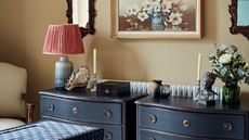 Two dark navy chest of drawers with candles, flowers, and lamps, against a beige wall. Above is a floral still-life and two wooden mirrors, with a patterned trunk and striped upholstered chair.