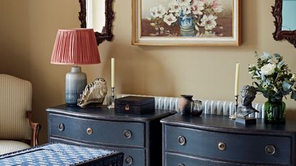 Two dark navy chest of drawers with candles, flowers, and lamps, against a beige wall. Above is a floral still-life and two wooden mirrors, with a patterned trunk and striped upholstered chair.
