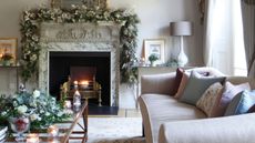 Neutral living room with marble fireplace and lit fire, pink couch, christmas garlands, glass coffee table, and lit candles.