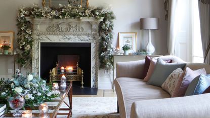 Neutral living room with marble fireplace and lit fire, pink couch, christmas garlands, glass coffee table, and lit candles.