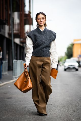 A woman walking in the street at Milan Fashion Week wearing tan pants, a white button down shirt, and gray sweater vest.