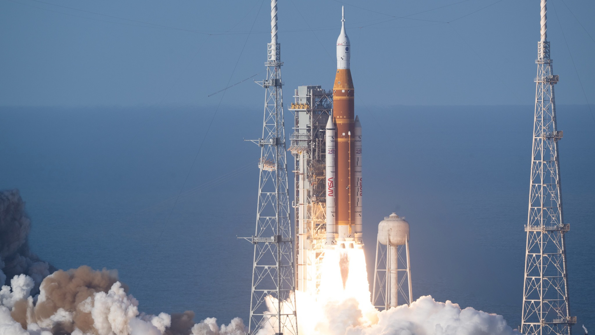 a huge orange and white rocket launches into a blue sky from a seaside pad