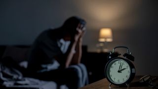 A man sitting on the edge of the bed with his head in his hands and a clock showing it's 2am