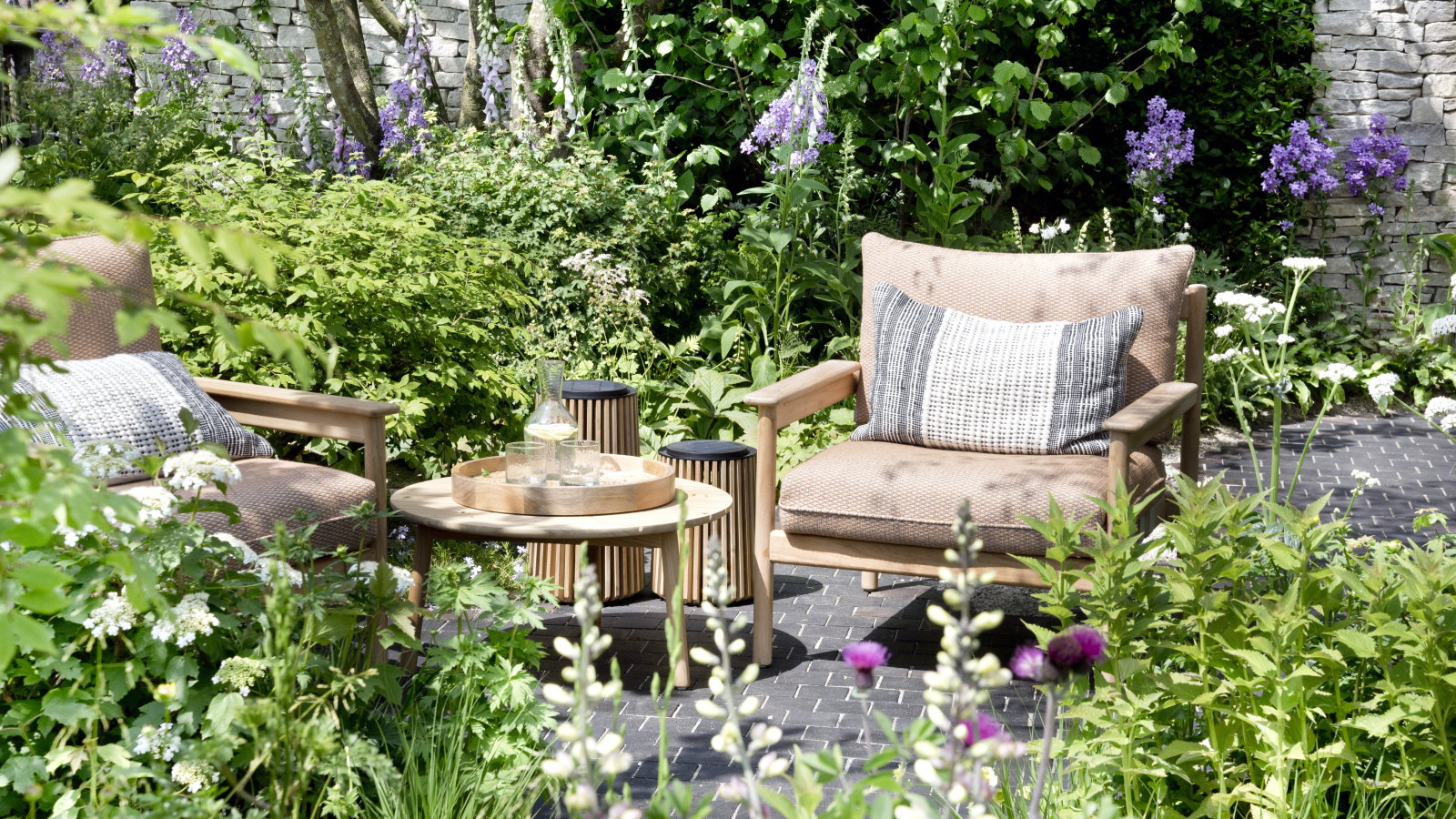 Packed flower beds around a paved seating area with a two chairs and a table