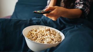 A bowl of popcorn in a bed with the hand of someone holding a TV controller