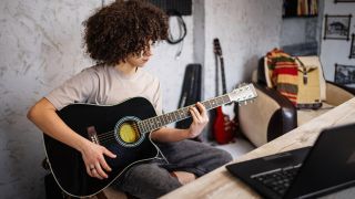 A boy plays acoustic guitar in front of a laptop