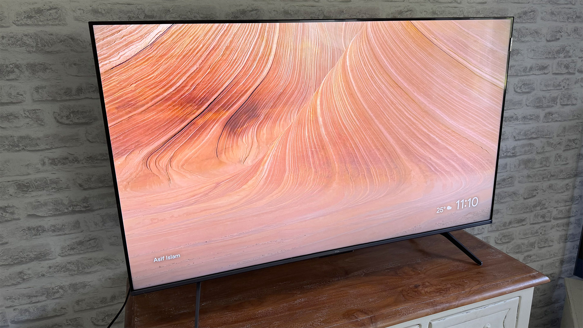 TCL C6KS 50-inch TV on wooden cabinet in front of brick wall, on screen is swirling desert pattern