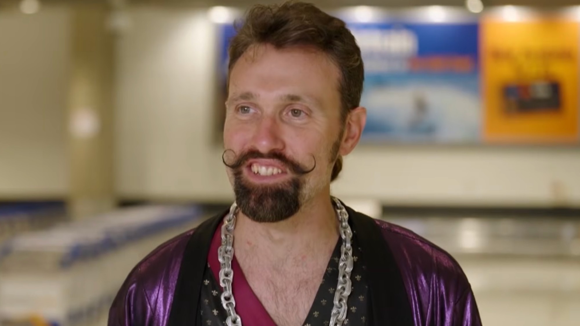 Nigel smiling with a waxed moustache, and he has a chain and purple shirt. 