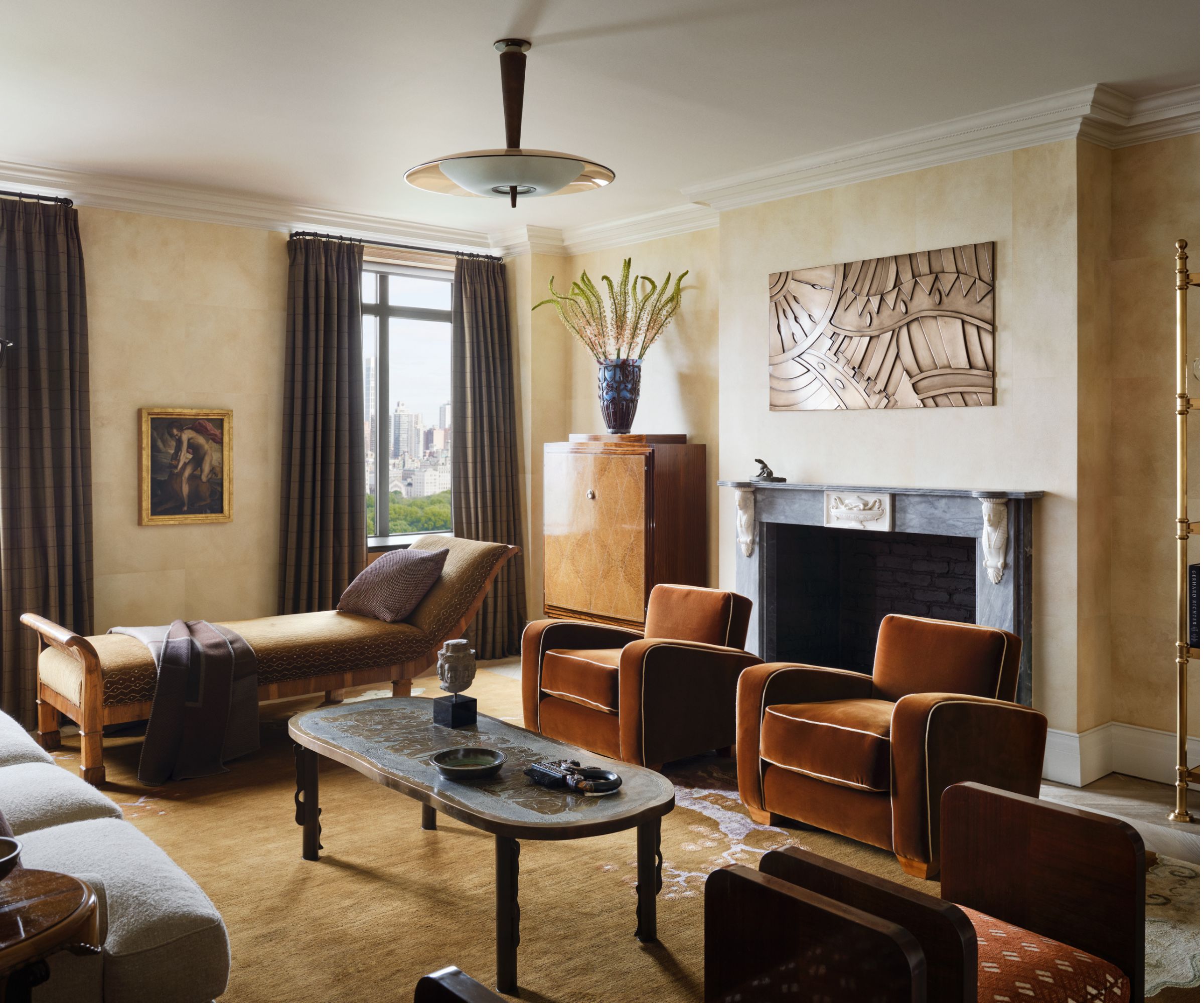 Art Deco style living room with velvet orange armchairs, a brown daybed, a marble coffee table, a mustard rug, a large fireplace, and a burl wood dresser in the corner.