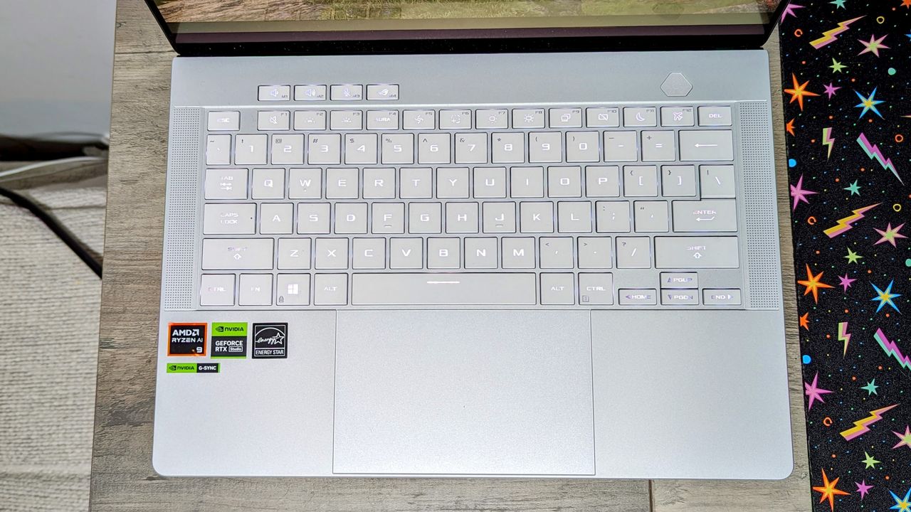 Asus Zephyrus G14 (2024) open on a brown table, highlighting the milky white keyboard and silver deck.
