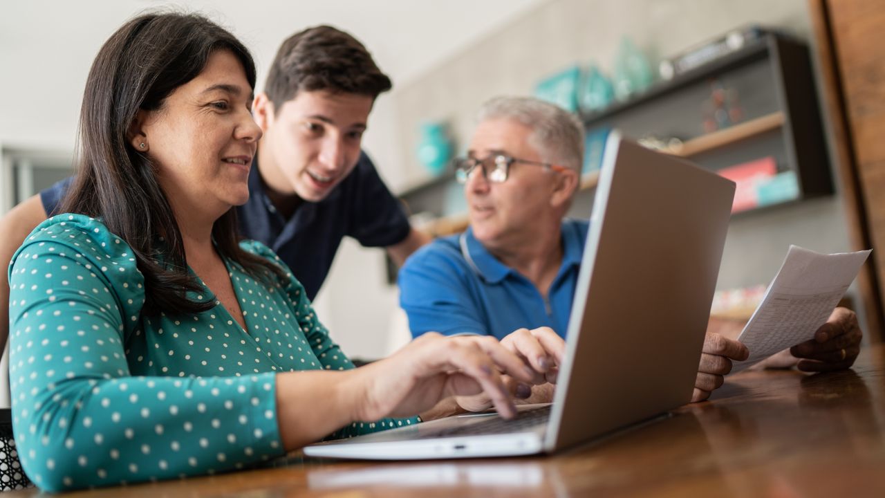 A mom works on a laptop while a teenage boy and his dad look on.