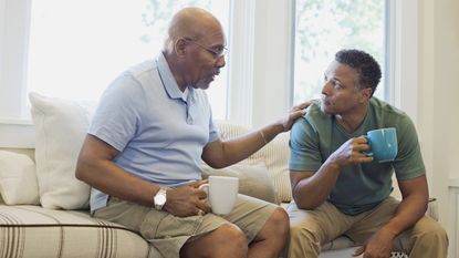 A father puts his hand on his son's shoulder as they talk while drinking coffee on the sofa.