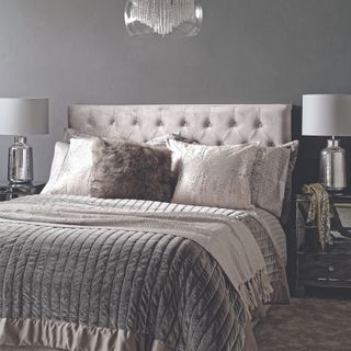 Grey and silver king sized bed with throw pillows and blanket, two bedside lamps eitherside in front of grey wall