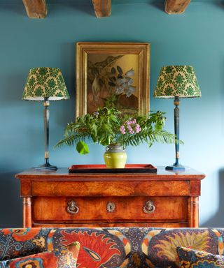 Blue living room with wooden sideboard, antique painting, two matching table lamps with green shades, vase of flowers and greenery, wooden ceiling beams, patterned upholstered seat