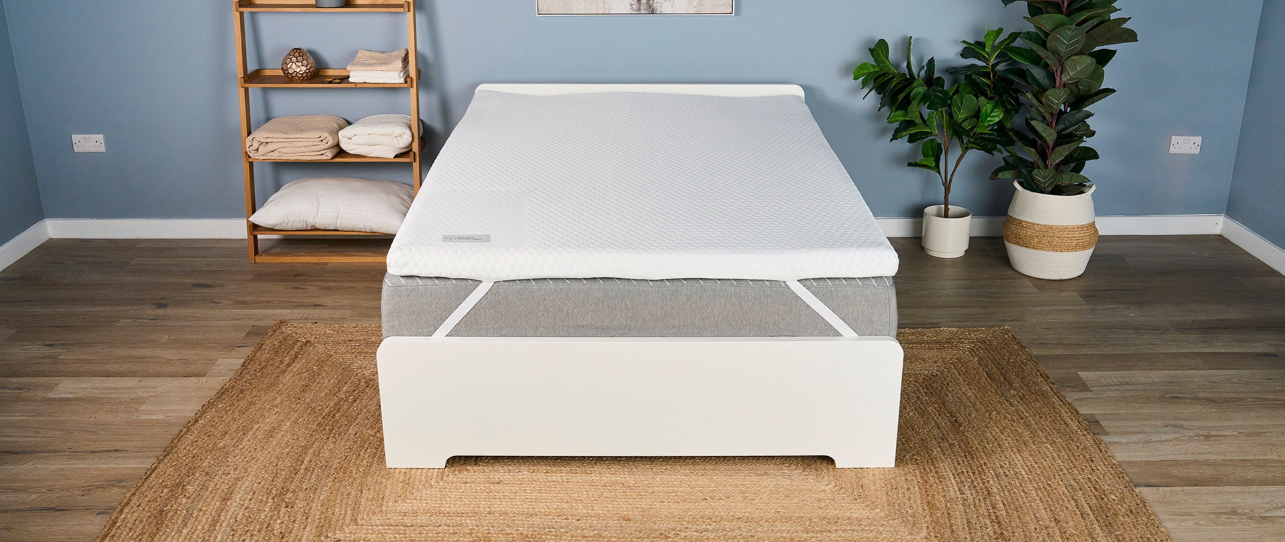 The Helix Dual Comfort Mattress Topper placed top of a grey bed on a white frame in a light blue bedroom with neutral-colored decor. 