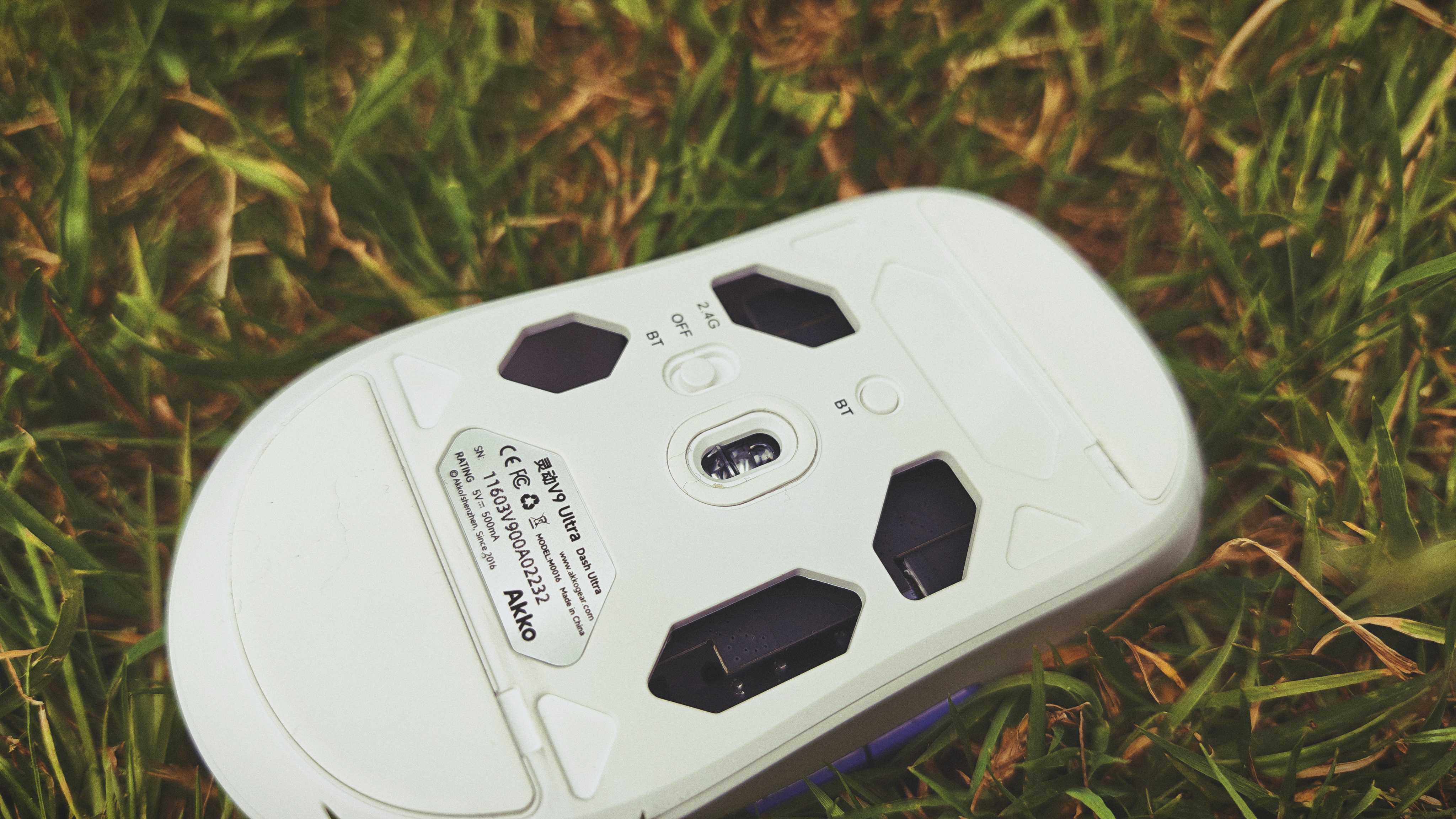 A white and purple Akko Dash Ultra gaming mouse sitting in tall grass in a garden