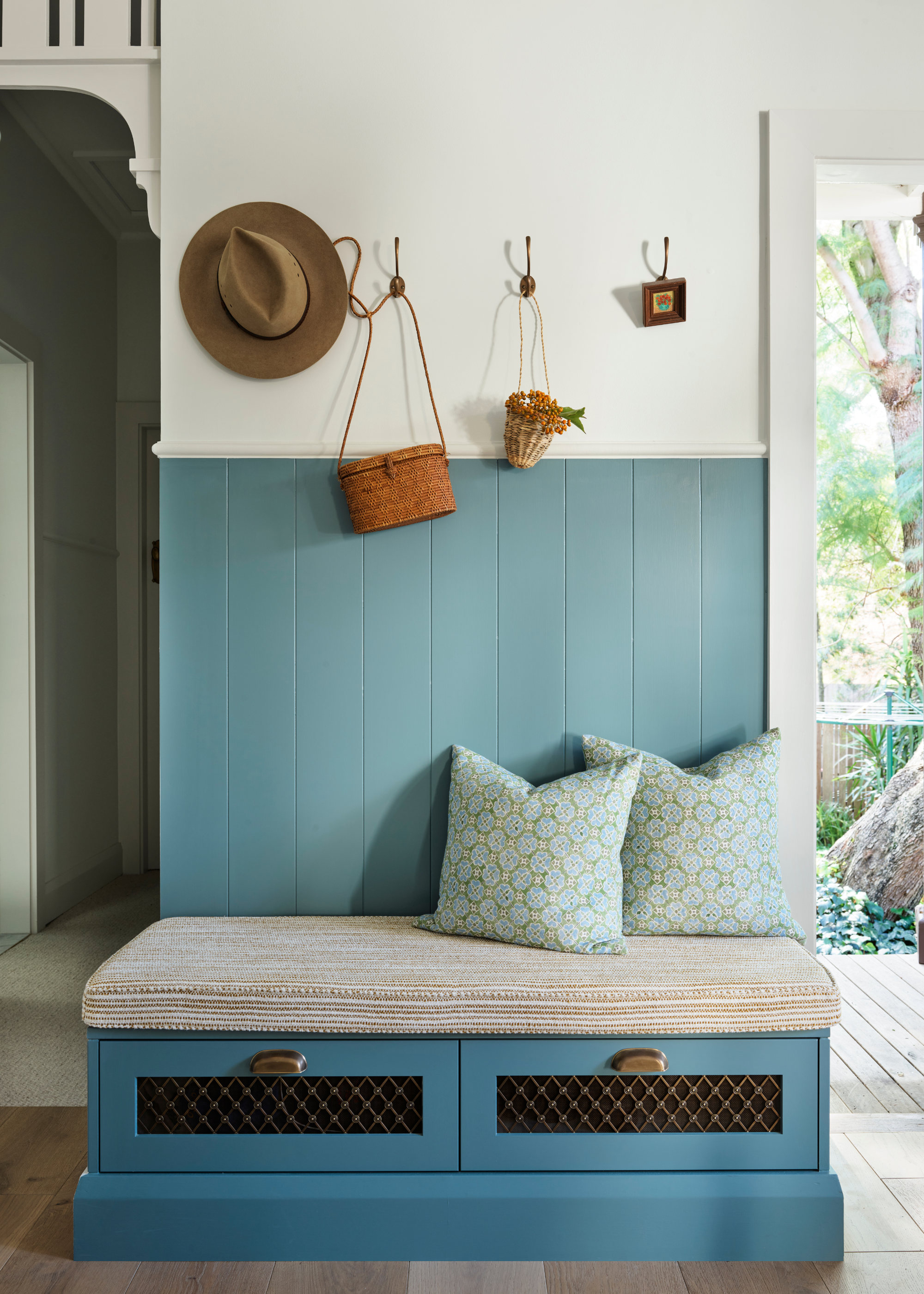 A mudroom with blue panelling and storage drawers along with a seat cushion and two pillows below a row of hooks with a hat and some bags