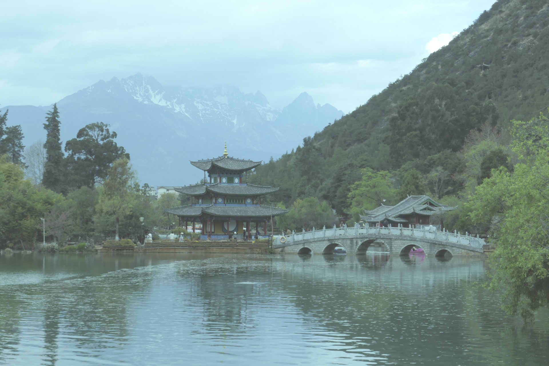 A Chinese temple by the side of a lake with an arched bridge leading up to it and both are reflected in the still water, in front of a snow covered mountain. A creative filter is applied to the image