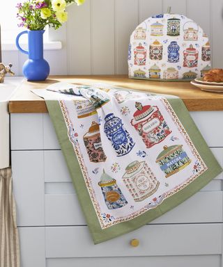 A kitchen towel with colorful tea tin illustrations with green borders hanging from a wooden counter with a blue vase of flowers and a tea cosy on it, and white drawers underneath it