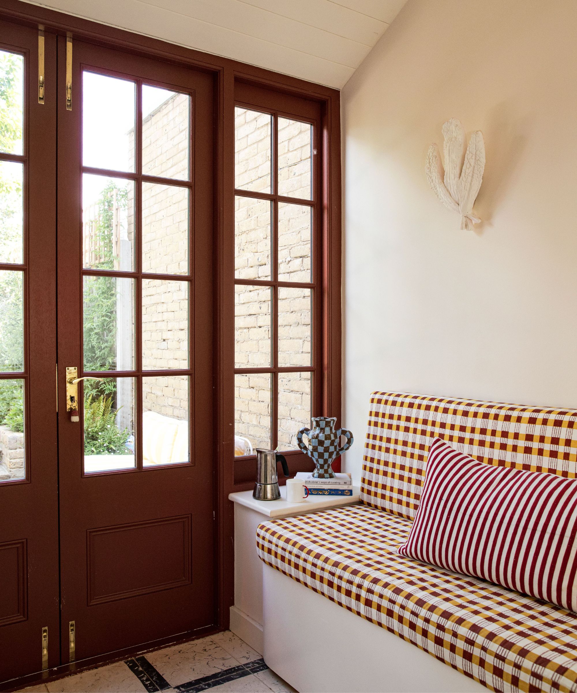 Conservatory with dark red glass doors, a built-in banquette with a geometric yellow cushion, and a side table holding a vase and a stack of books.