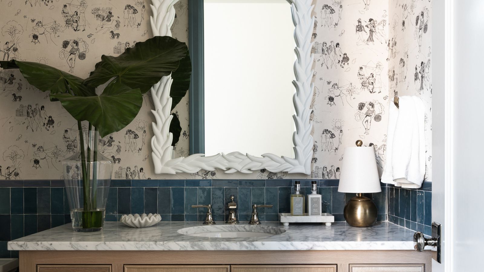 A close-up of a wooden bathroom vanity with a marble countertop decorated with a vase of leaves and a gold lam