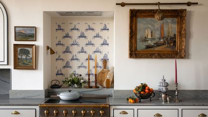 Vintage kitchen with white walls, an alcove with Delft tiles, a range oven tucked in, an antique painting of a seascape, antique candlesticks and a fruit bowl beneath.