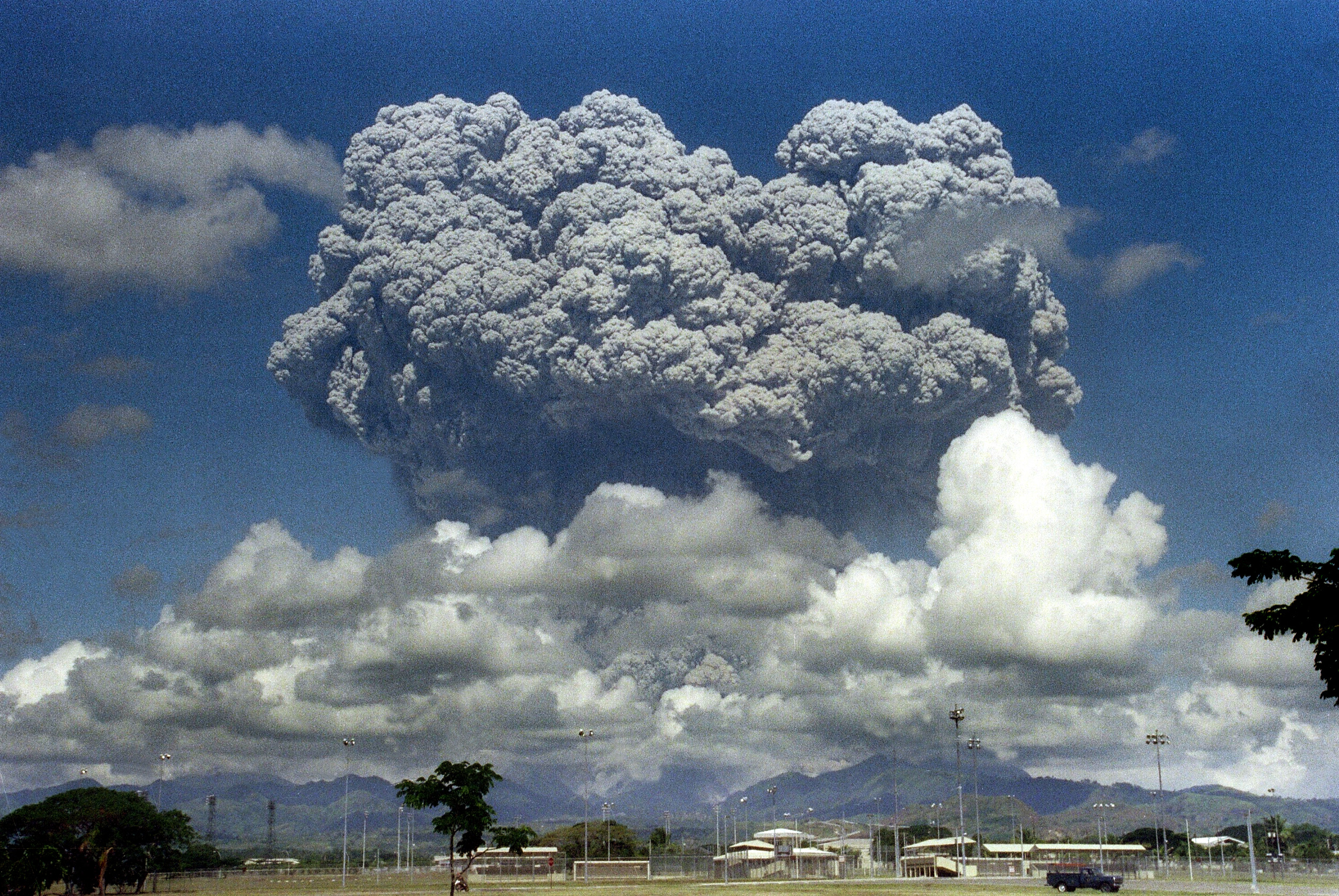 a big mushroom cloud from a volcano erupting with fields and buildings in foreground