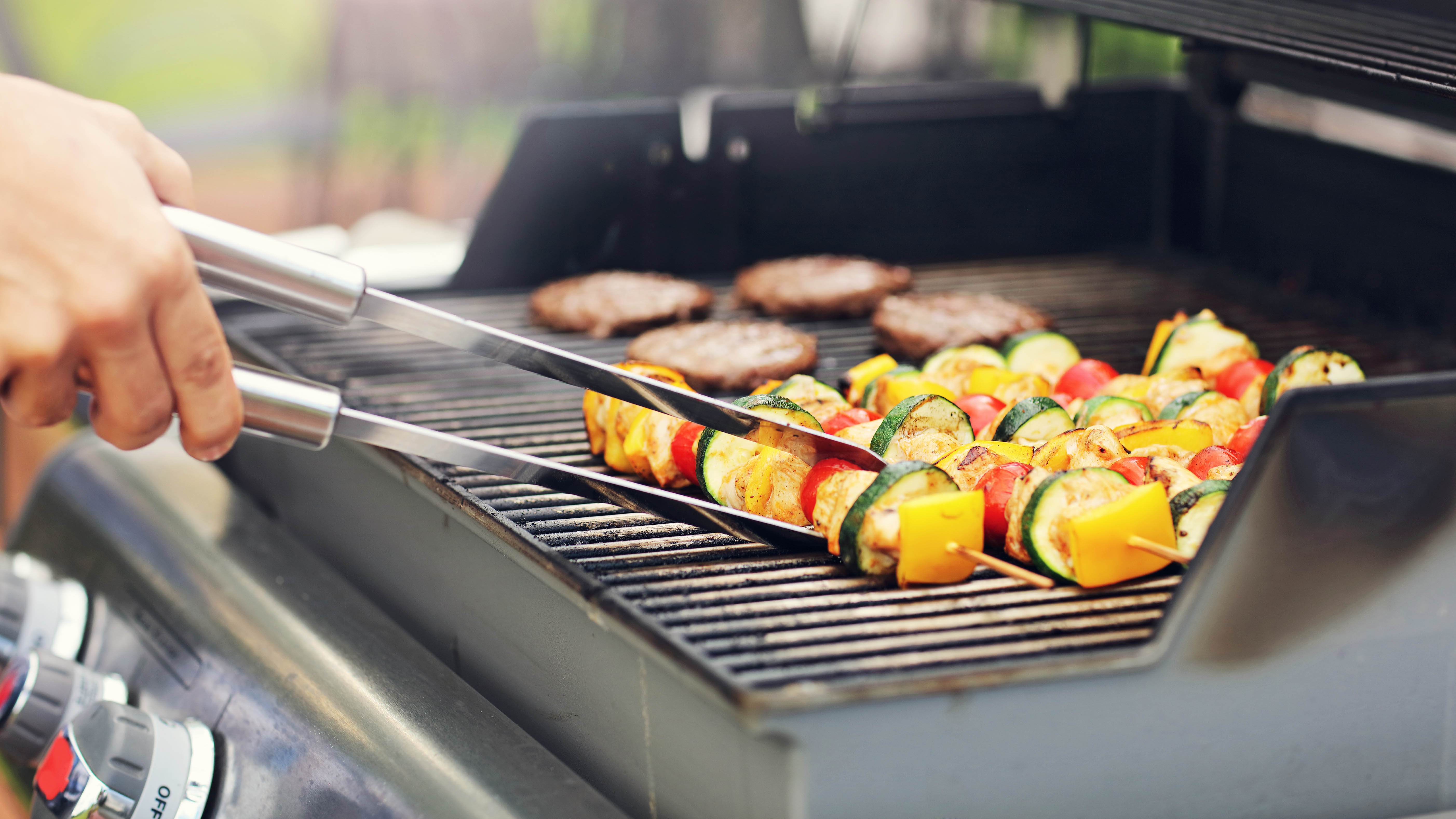 Someone grilling vegetable skewers and burgers on a gas grill