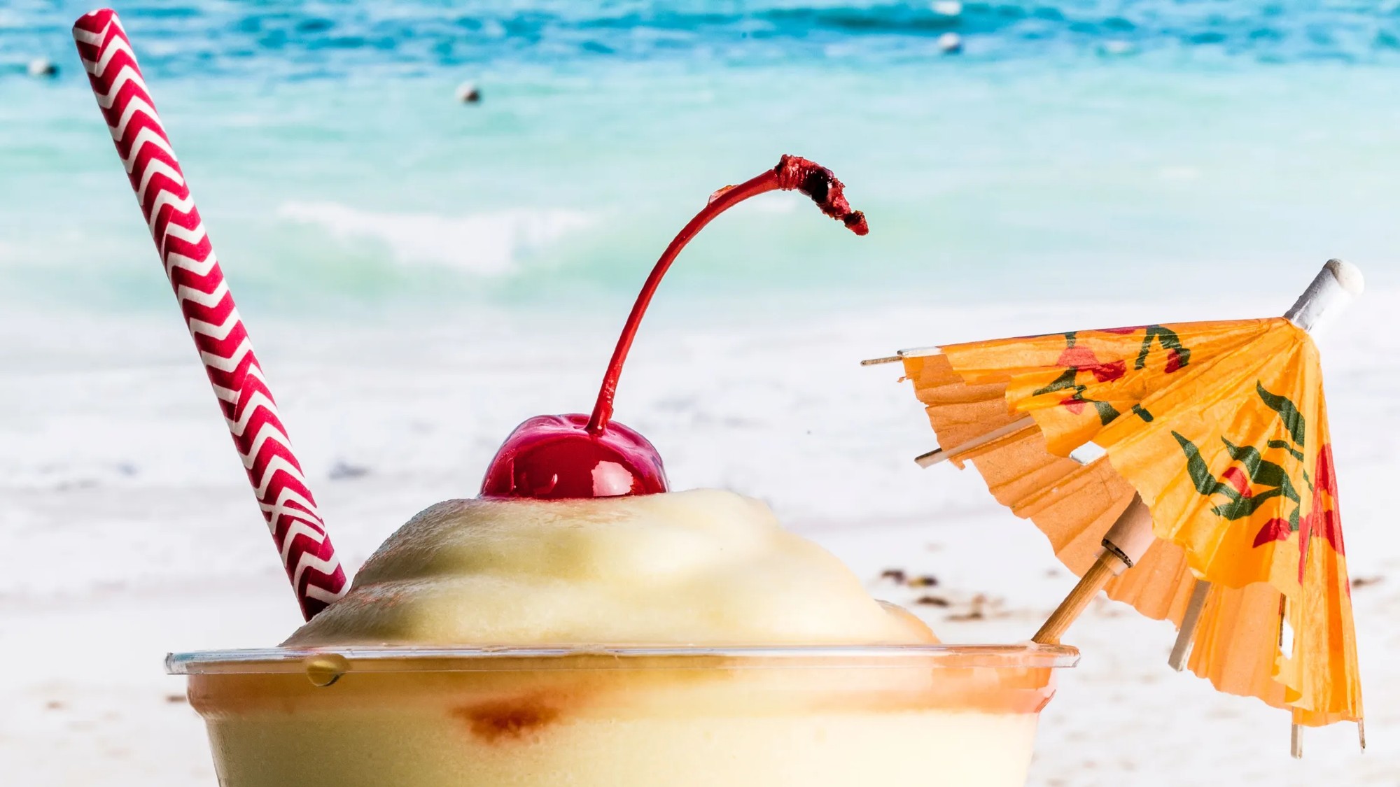 side shot of the top of a piña colada, with a paper-umbrella and maraschino cherry garnish