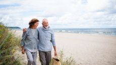 Happy senior woman and man embracing and walking outdoors on sandy beach in autumn.