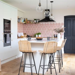 white kitchen with pink tiled splashback with kitchen island and woven wicker bar stools