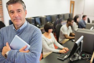 Male teacher in blue sweater stands in classroom with teens on computers in the background.