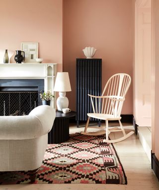 blush pink living room with black tall boy, white painted floor, pale gray couch, fire place, windsor chair, white accessories, patterned rug,