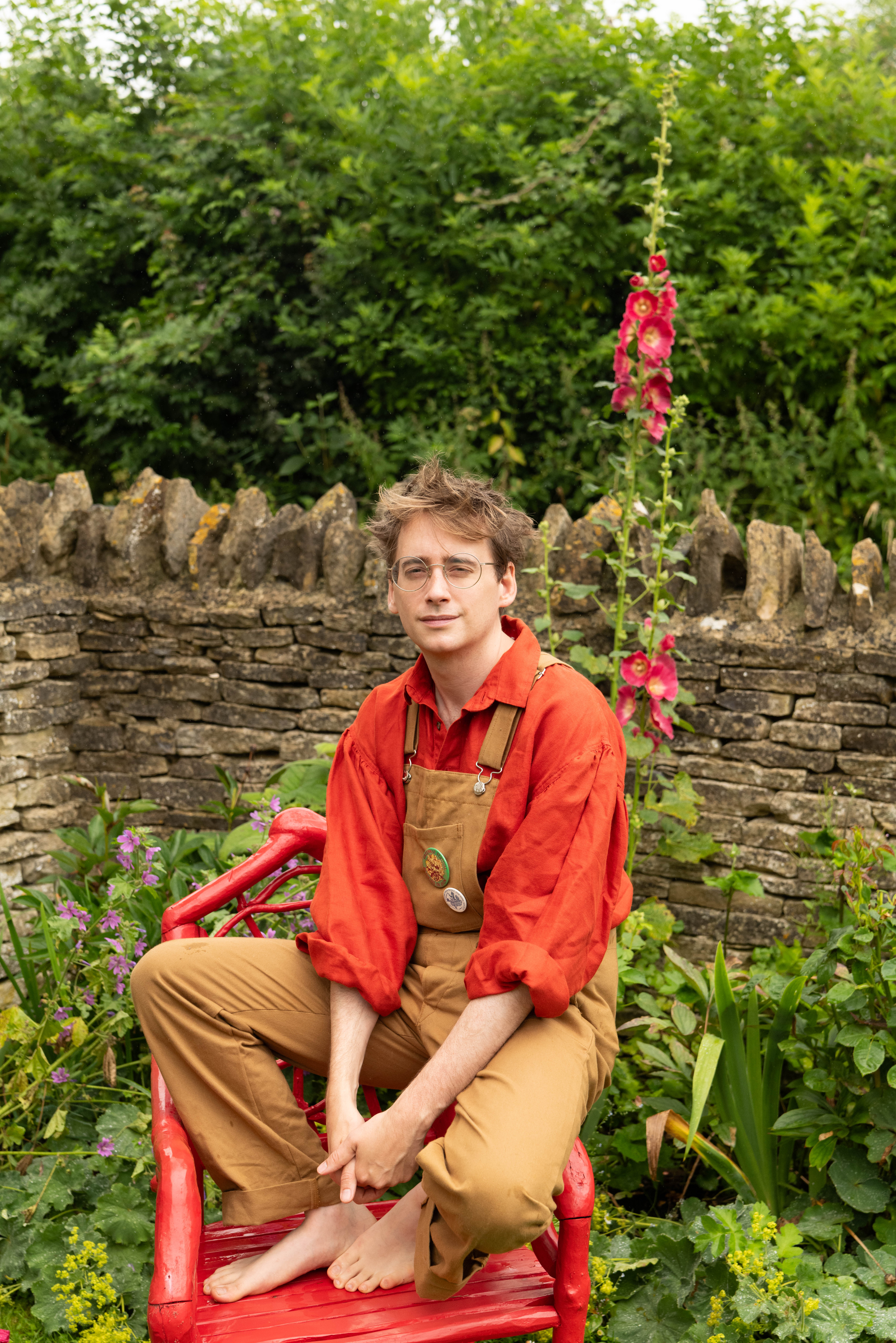 Man in dungarees perched on a red chair in a garden